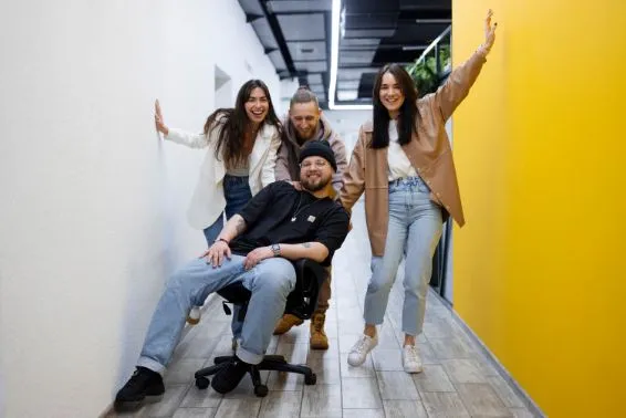 Fünf junge Menschen in einem Büroflur, zwei stehen an der Wand, einer sitzt auf einem Bürostuhl und zwei stehen hinter ihm. Sie tragen alle legere Kleidung.